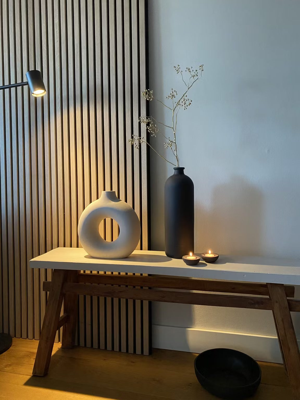 Decorazione minimalista con un vaso circolare bianco, un vaso alto nero con steli secchi, due candele tealight e una ciotola nera su uno sfondo acustico a doghe di legno Akupanel.